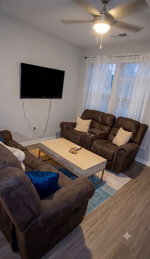 Un salon doté d’un téléviseur à écran plat fixé au mur, d’un ventilateur de plafond, d’un canapé en cuir marron, d’une table basse en bois et d’un tapis à motifs.