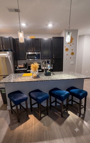 Una cocina moderna con muebles oscuros, un microondas de acero inoxidable y una encimera de granito. Hay tres taburetes azules y un jarrón decorativo con flores amarillas.