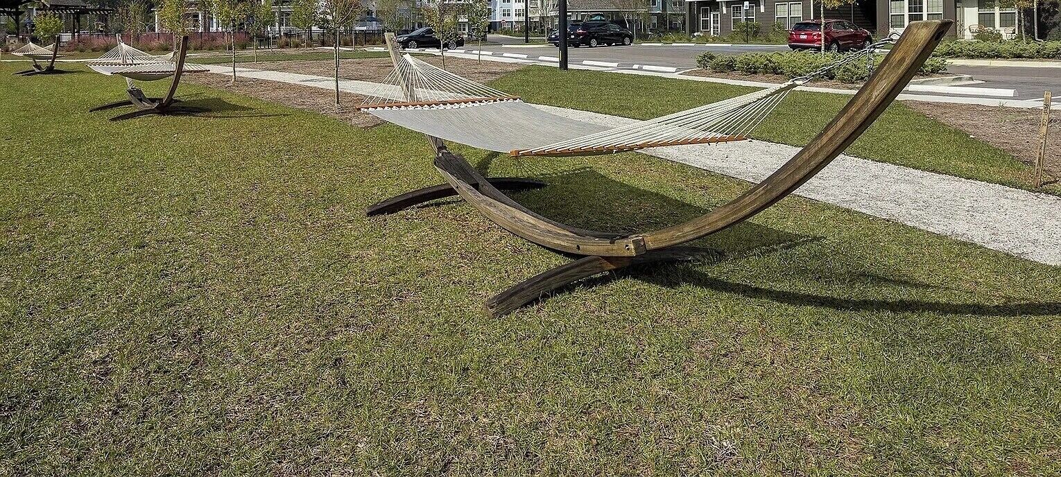 A hammock-style seating area with a metal frame and a flat surface, surrounded by grass and a paved walkway.