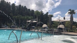 En swimmingpool med klart vand, omgivet af liggestole og parasoller, med et baggrundslandskab af træer og en delvist overskyet himmel.