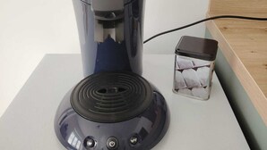Cafetera con depósito de agua y cable de alimentación sobre una superficie blanca.