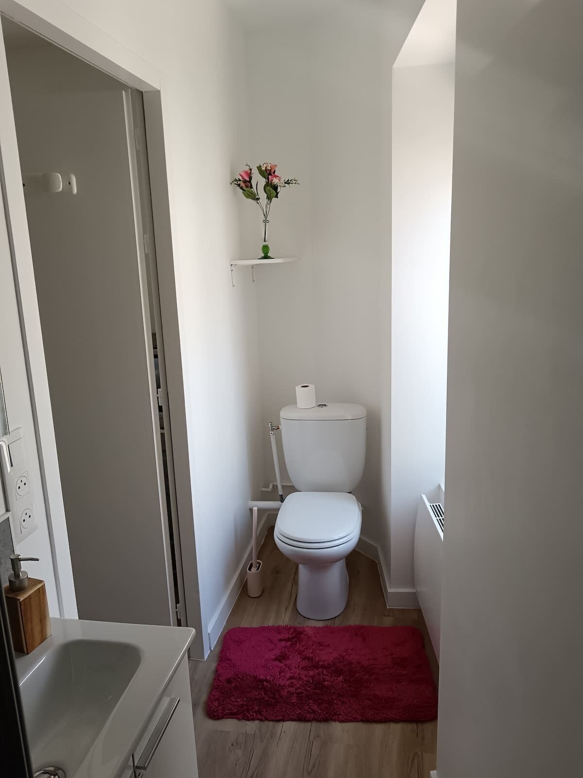 Ein kleines Badezimmer mit Toilette, einer kleinen Ablage mit einer Vase, einem roten Teppich und Holzboden.