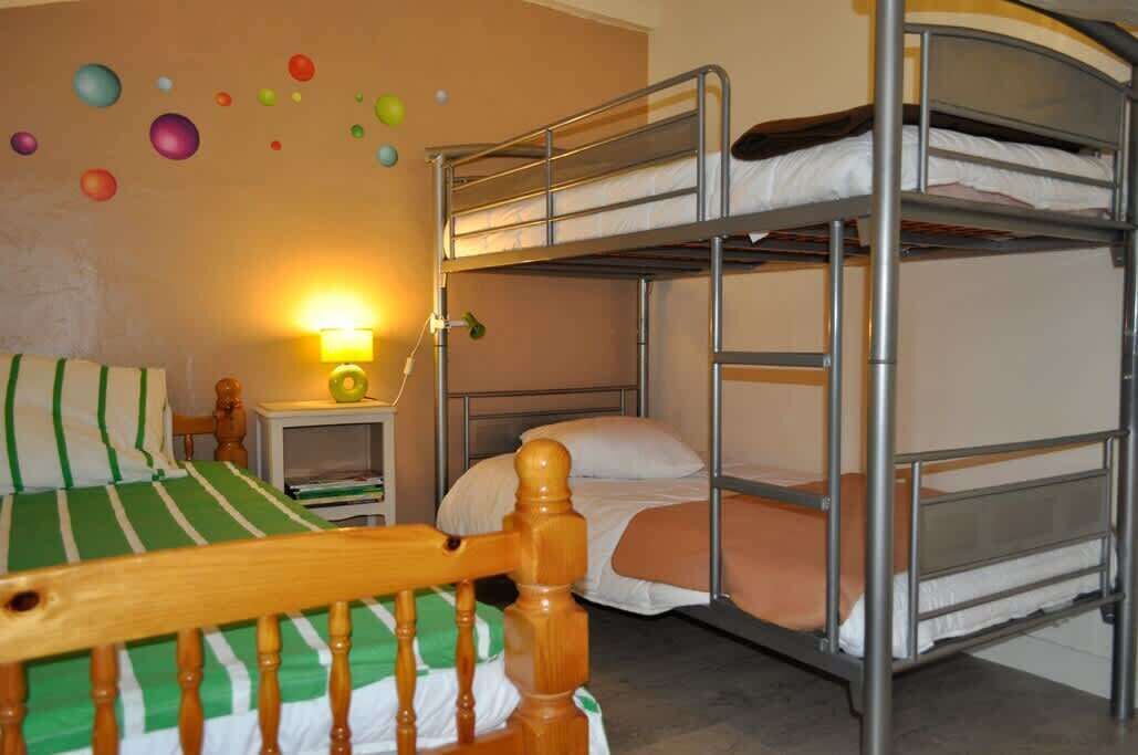 Habitación con literas, una silla de madera, una mesita de noche con lámpara y decoraciones de pared coloridas.