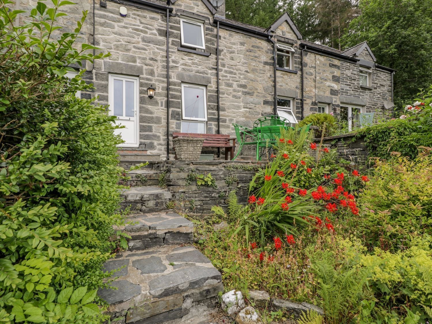 A stone building with a stone staircase leading to a door, surrounded by lush greenery and red flowers.