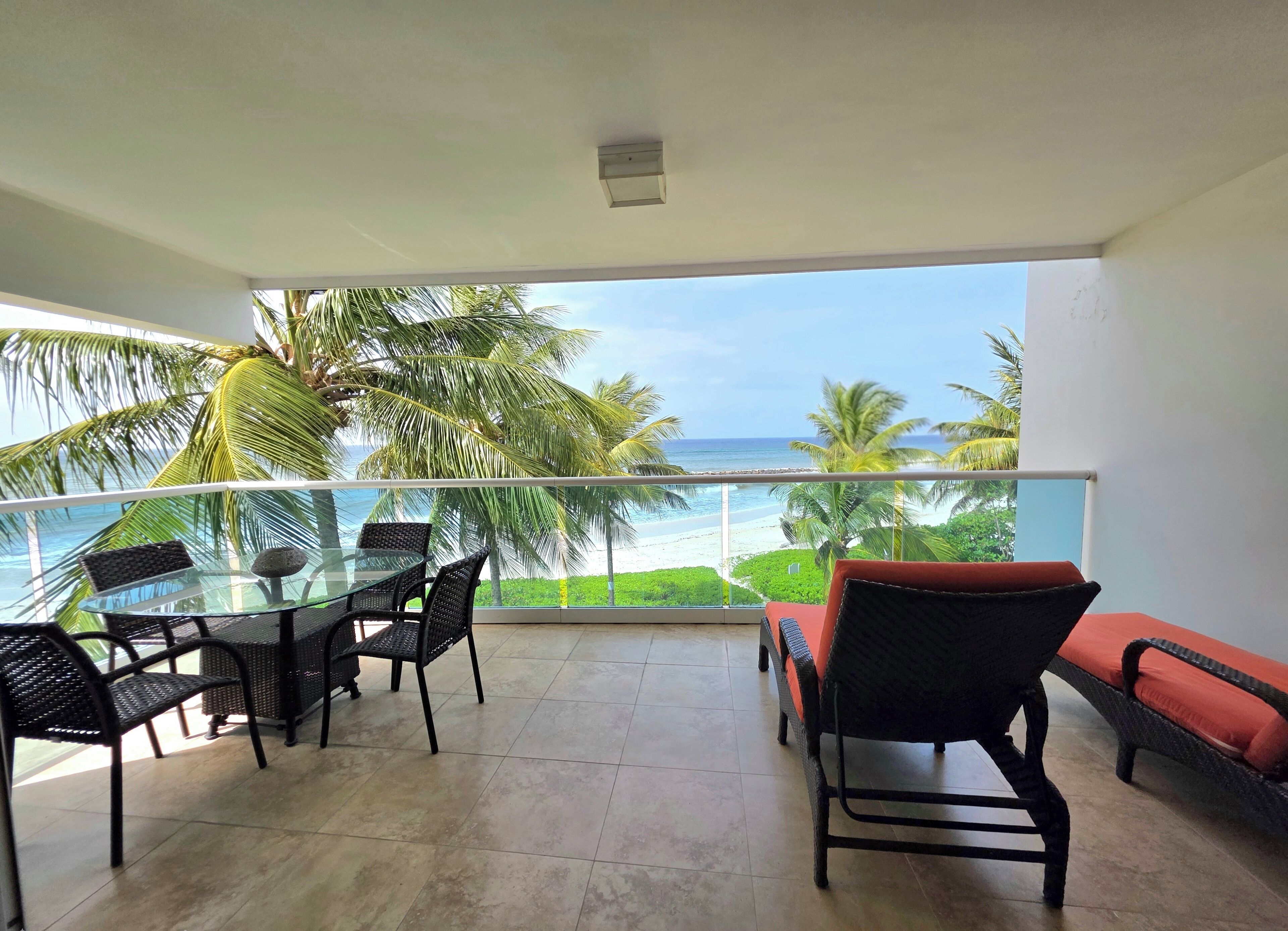 Un balcón con una mesa y sillas de vidrio, con vista a una playa y palmeras.