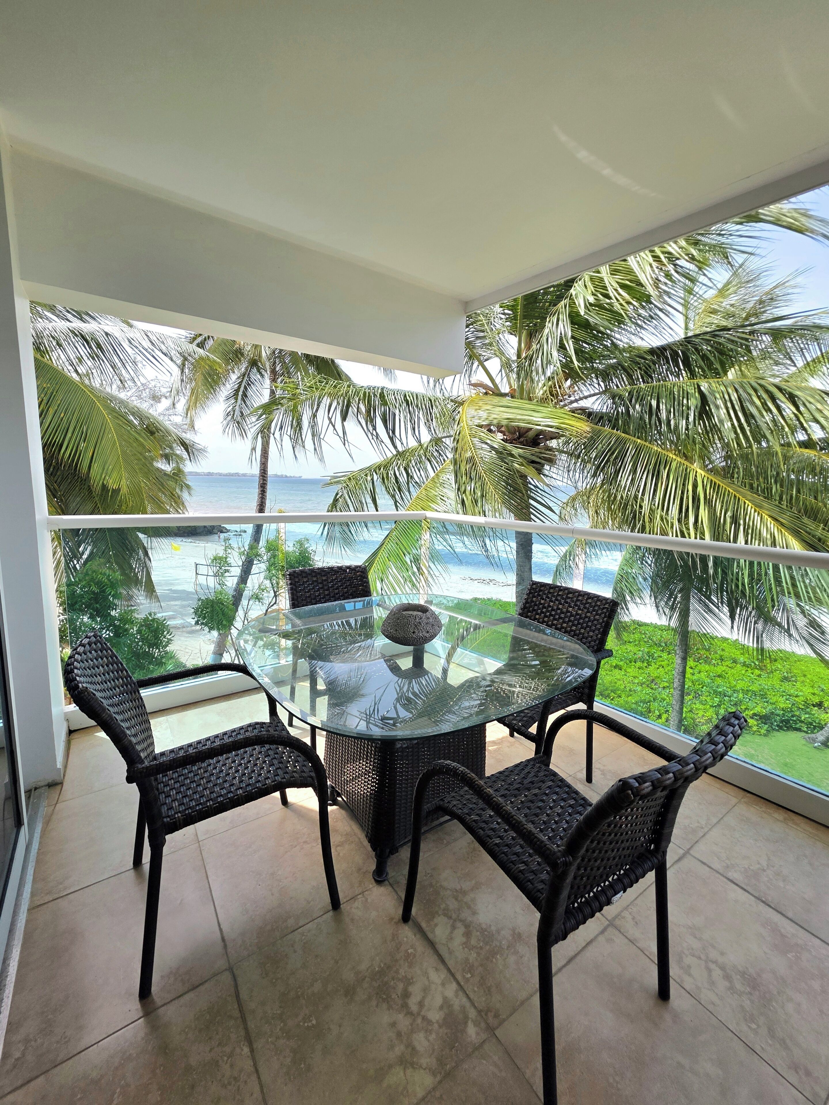 A balcony with a glass table and wicker chairs, overlooking a beach and palm trees.