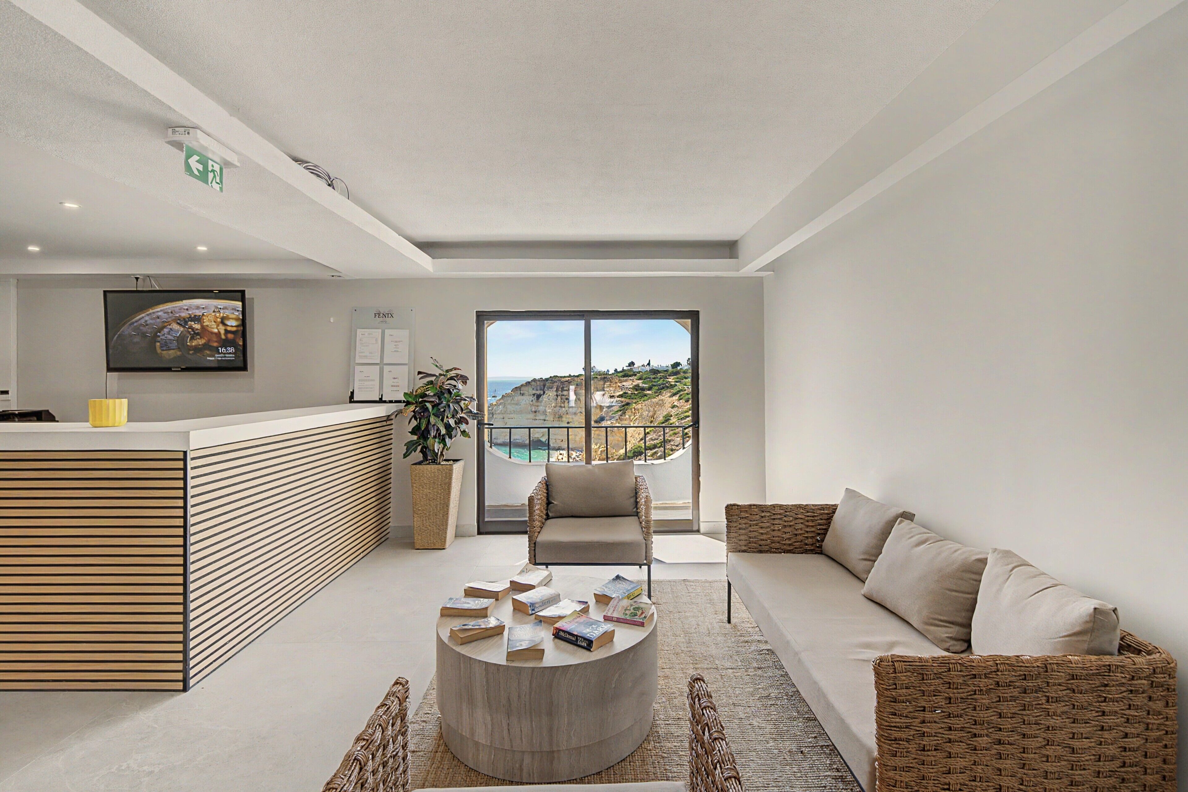 A modern hotel lobby with a reception desk, a sofa, a coffee table with books, a TV, and a large window with a view of cliffs.