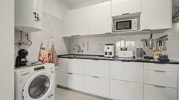 Una cocina moderna con armarios blancos, microondas integrado, cafetera y lavadora.