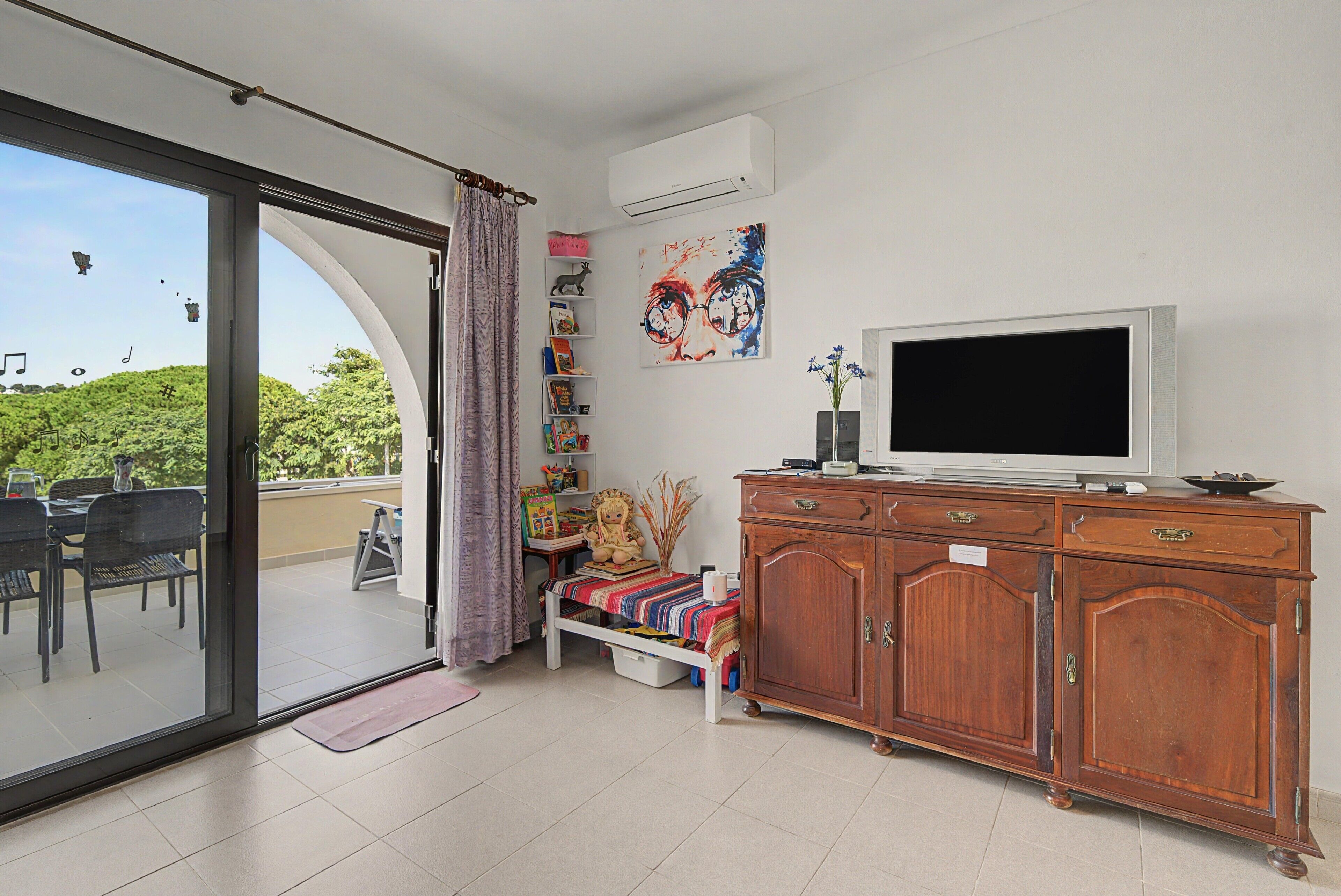Un salón con puerta corrediza de vidrio que da a un balcón, un televisor, una estantería y un mueble de madera.