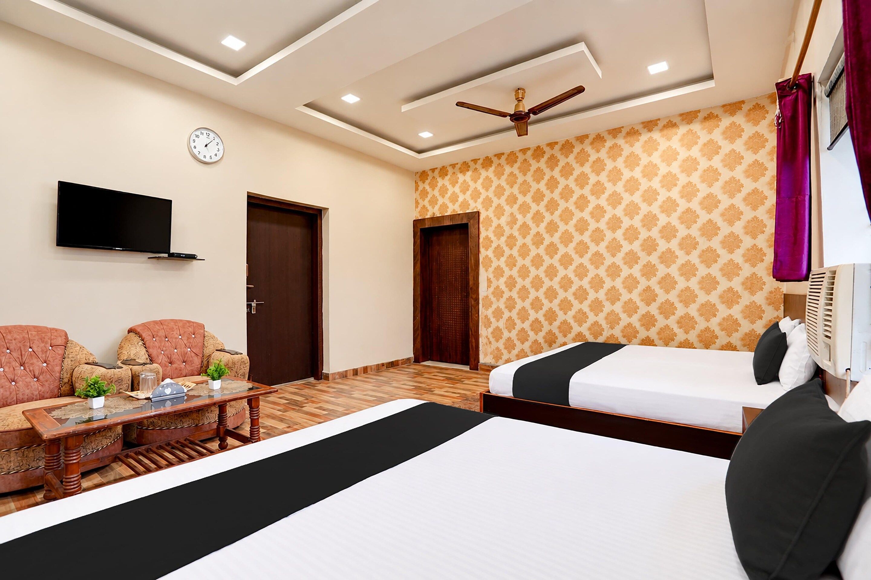 A hotel room with a bed, two armchairs, a small table, a TV, a ceiling fan, and patterned wallpaper.