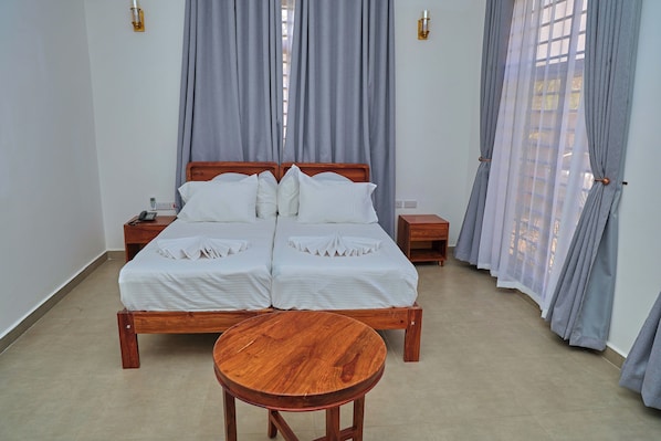 Una cama doble con ropa de cama blanca, dos mesitas de noche, una mesita auxiliar de madera y una mesa redonda de madera en una habitación con cortinas grises y suelo de baldosas.