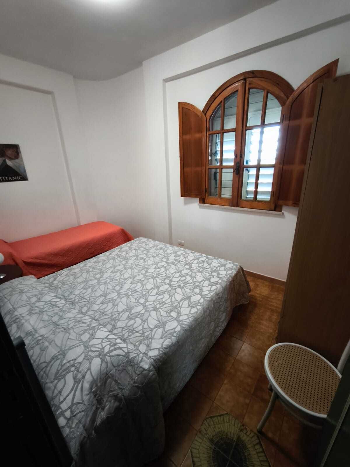 A bedroom with a bed, a chair, a window with wooden shutters, and a poster on the wall.