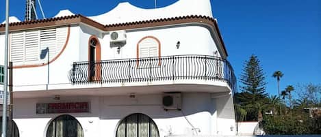 A two-story building with a white facade, a balcony with a black railing, and a sign that reads 'FARMACIA LA MEDITERRANEA'.