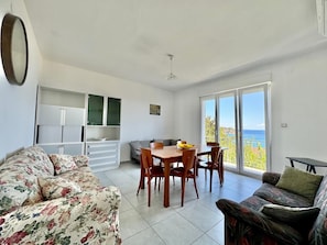 A room with a dining table, chairs, a sofa, and a balcony with a view.