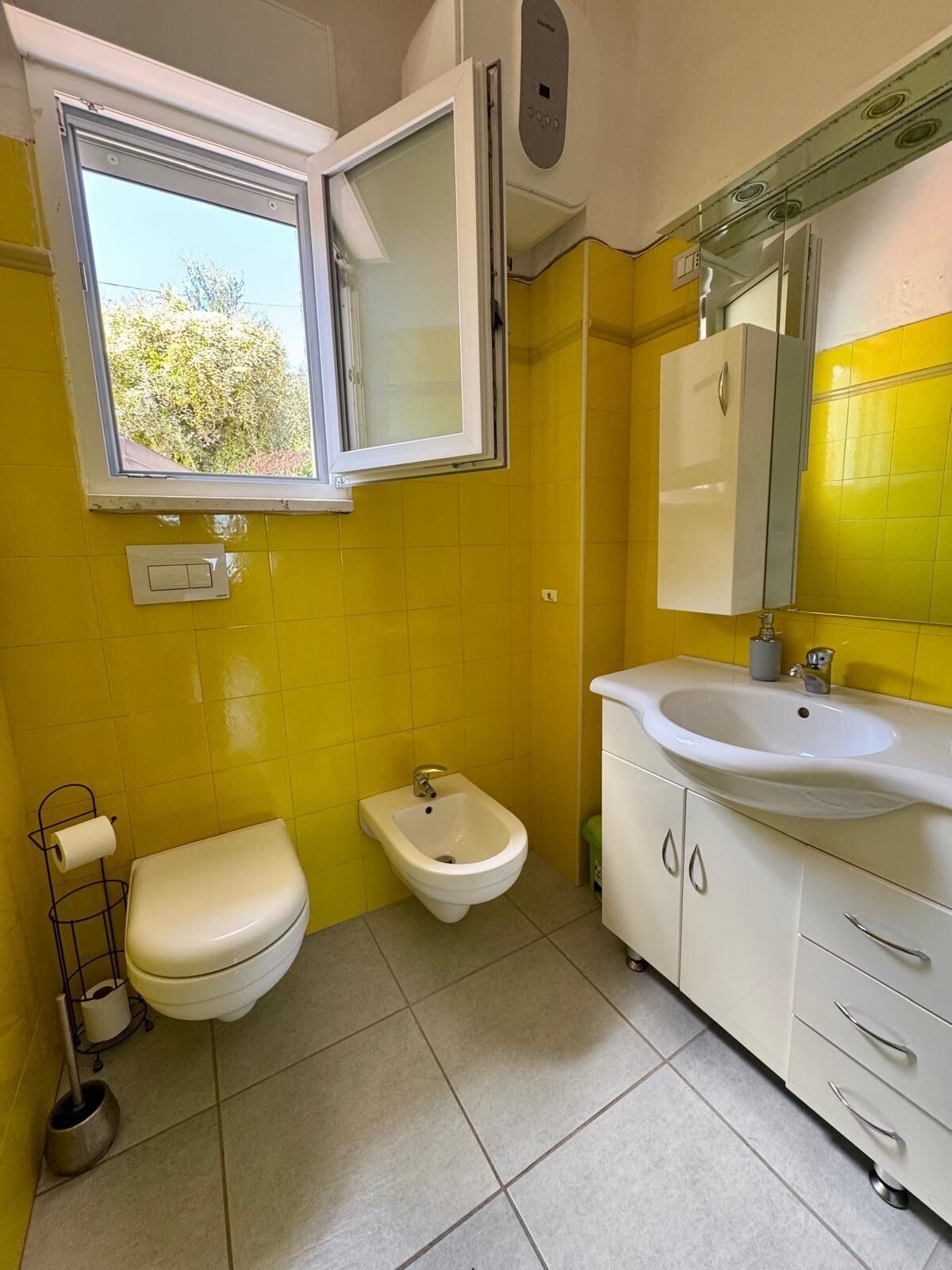 Ein Badezimmer mit gelben Wänden, einer weißen Toilette, einem Bidet, einem Waschbecken mit Spiegel und einem Fenster mit Blick auf Bäume.