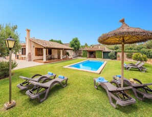 A poolside area with lounge chairs, a thatched umbrella, and a house with a tiled roof. - Family Villa Between Pollenca Towns (Pollença)