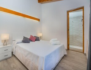 A bedroom with a bed, two bedside tables, a nightstand, a wooden ceiling beam, a door, and a view of the beach. - Family Villa Between Pollenca Towns (Pollença)