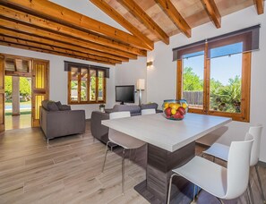 A modern living room with a wooden ceiling, a dining table with chairs, a television, and a bowl of fruit. - Family Villa Between Pollenca Towns (Pollença)