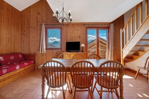 A wooden interior with a dining table, chairs, a sofa, a TV, and a staircase.