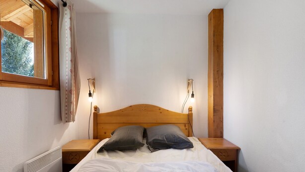 A bedroom with a wooden headboard, two bedside lamps, a radiator, and a window with curtains. - Le 8D des Cimes La Toussuire (Villarembert)