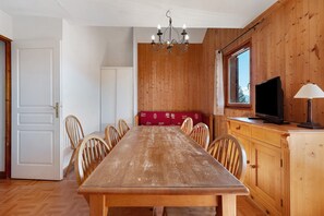 A dining area with a wooden table and chairs, a chandelier, a television on a wooden cabinet, and a window with a view of trees.