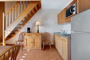 A kitchen with wooden cabinets, a built-in microwave, a refrigerator, a coffee maker, and a wooden chair. - Le 8D des Cimes La Toussuire (Villarembert)