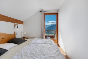 A bedroom with a bed, a nightstand, a lamp, a window with a mountain view, and a wooden ceiling beam. - Le 8D des Cimes La Toussuire (Villarembert)