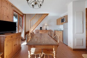A dining area with a wooden table and chairs, a television on a cabinet, and a kitchen with a microwave and refrigerator. - Le 8D des Cimes La Toussuire (Villarembert)