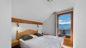 A bedroom with a wooden headboard, a bed with white bedding, a nightstand, a lamp, and a view of mountains through a balcony door.