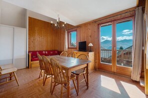 A wooden interior with a dining table, chairs, a television, and a view of mountains through large windows.