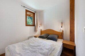 A bedroom with a wooden headboard, a single bed with white bedding, a nightstand, a wall-mounted lamp, and a window with a view of greenery. - Le 8D des Cimes La Toussuire (Villarembert)