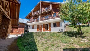 A two-story wooden chalet with a balcony on the upper floor, a sloped roof, and a small garden area.