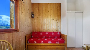 Interior de fusta amb un llit sofà, una cadira de fusta, una taula i vista a una muntanya a través d'una finestra.
