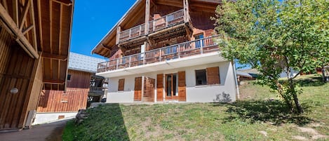 A two-story wooden chalet with a balcony on the upper floor, a sloped roof, and a small garden area.