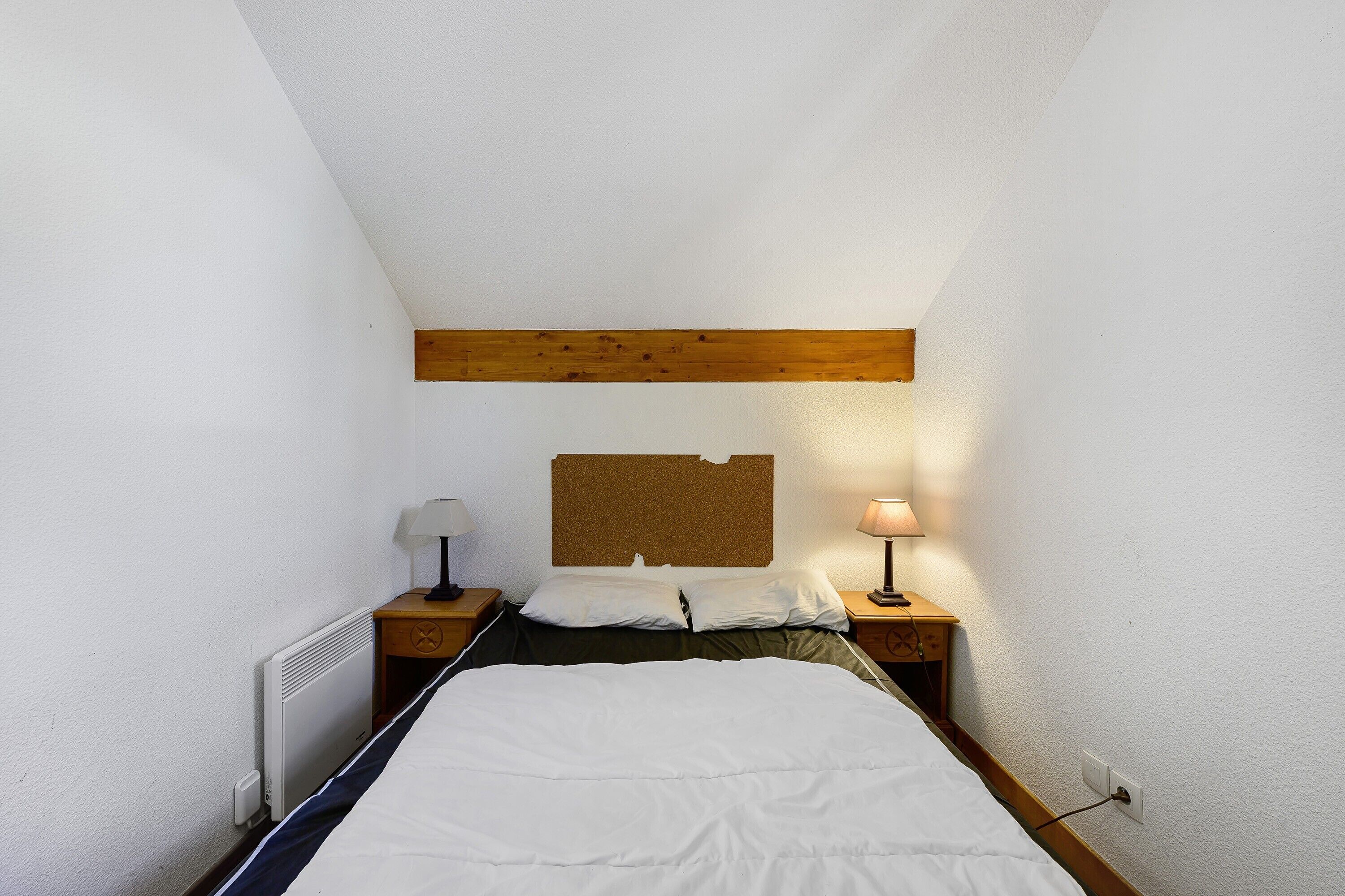 A small, single-bed room with a sloped ceiling, a wooden beam above the bed, a cork bulletin board, two bedside lamps, and a radiator.