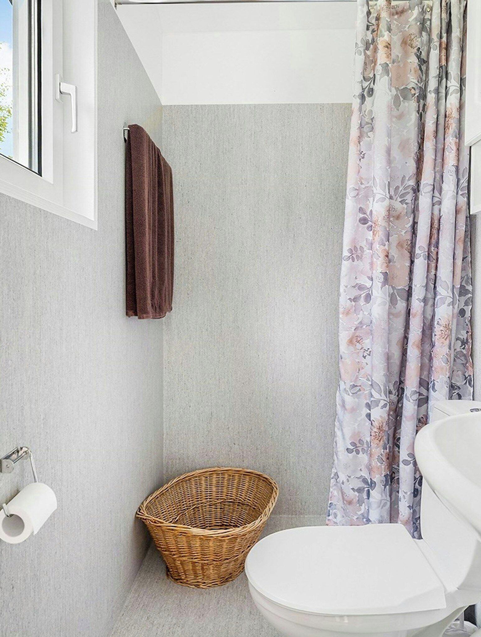 A bathroom with a white toilet, a wicker basket, a brown towel, and a floral shower curtain.