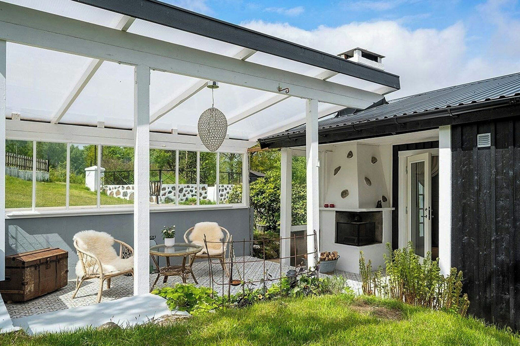 A covered outdoor seating area with wicker chairs and a table, a grill, and a view of a garden and house.