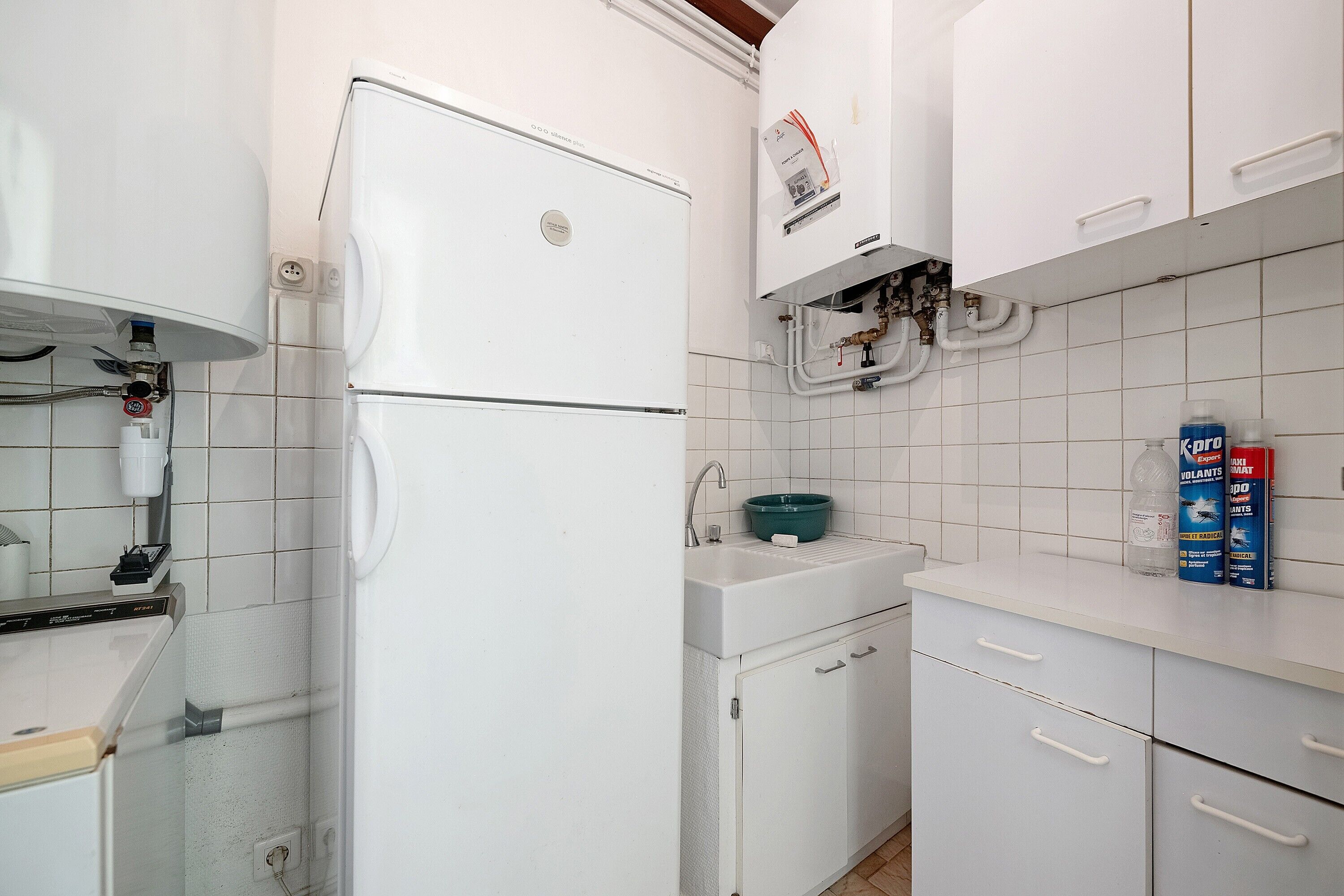 Una cocina con armarios blancos, un refrigerador blanco, un lavamanos con lavabo verde y un calentador de agua.