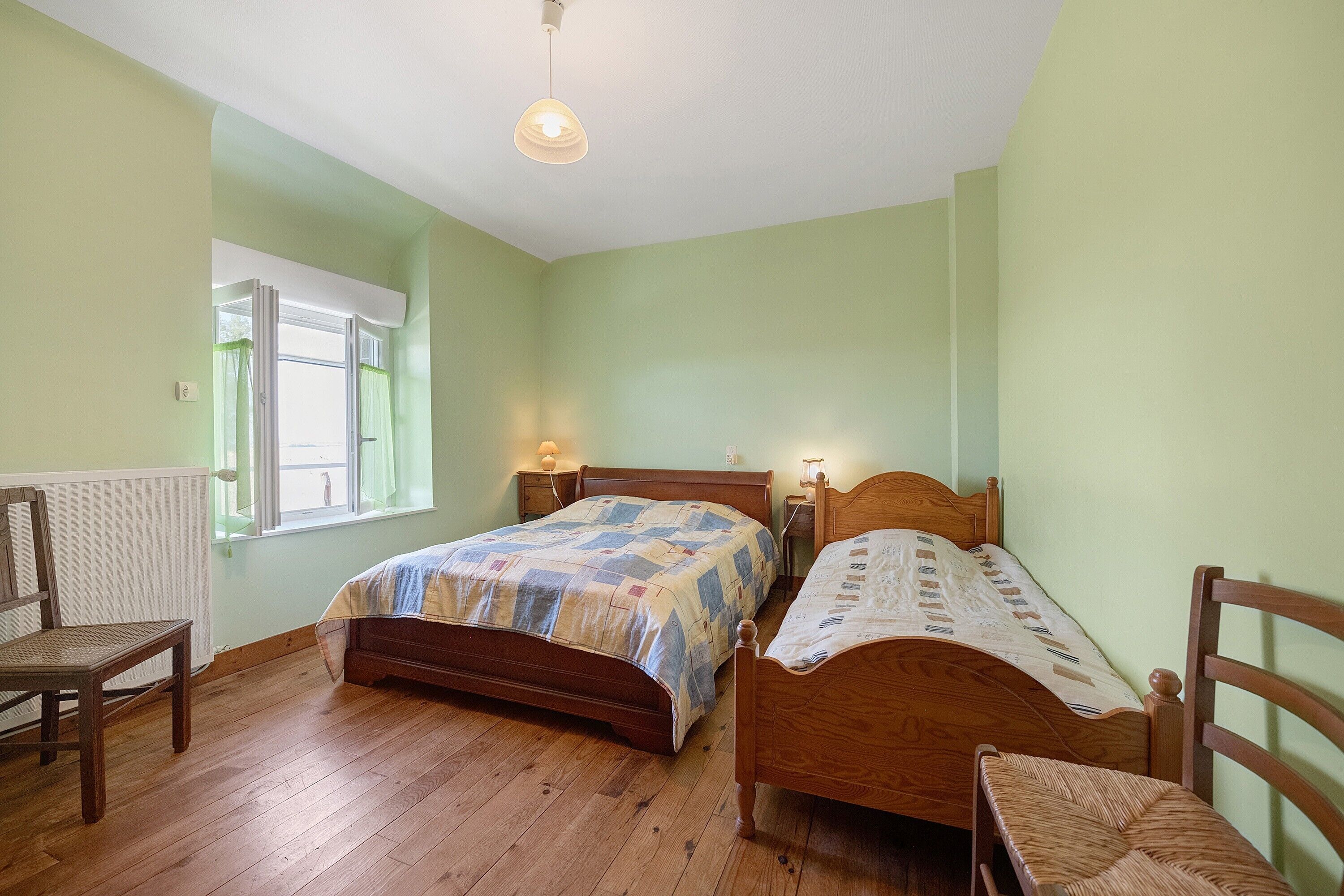 A bedroom with two wooden beds, a chair, a window with curtains, and a hanging light fixture.