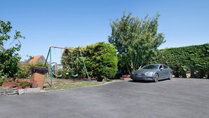 Une allée menant à une maison, avec une voiture garée, un portique et un jardin agrémenté de plantes en pot.