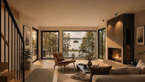Uma sala de estar moderna com lareira, um sofá, uma poltrona e vista para um lago através de grandes janelas.
