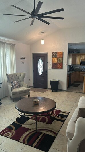A living room with a round coffee table, a sofa, a chair, a ceiling fan, a door with a glass panel, and a kitchen area visible through an open door. - Charming 2-bedroom house with AC in sunny Orlando (Orlando)