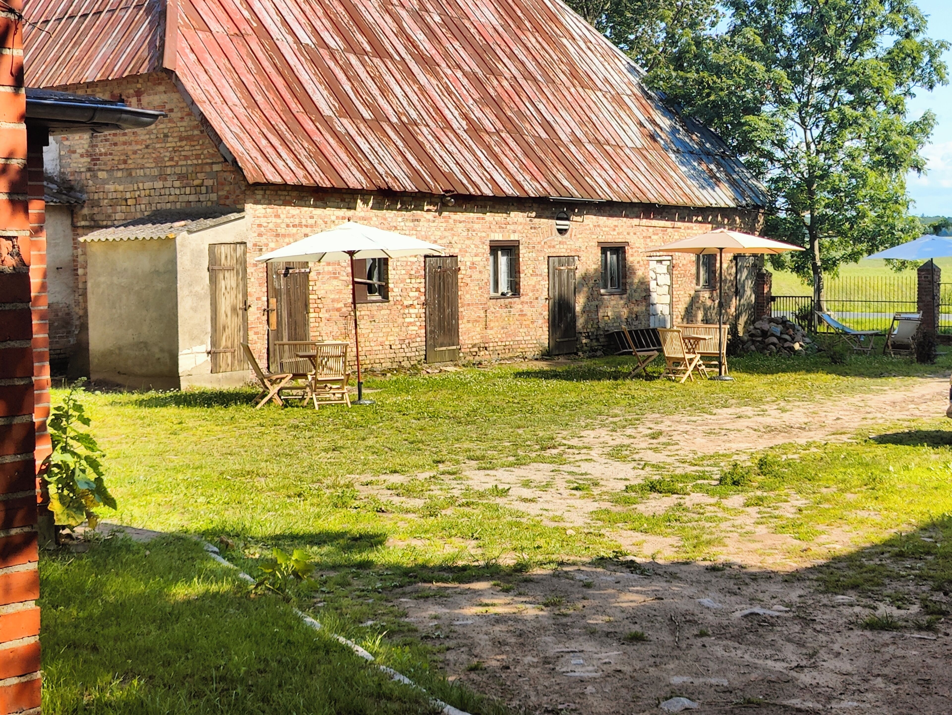 A rustic brick building with a corrugated metal roof, a small outdoor seating area with wooden chairs and tables, and a dirt path leading to the entrance.