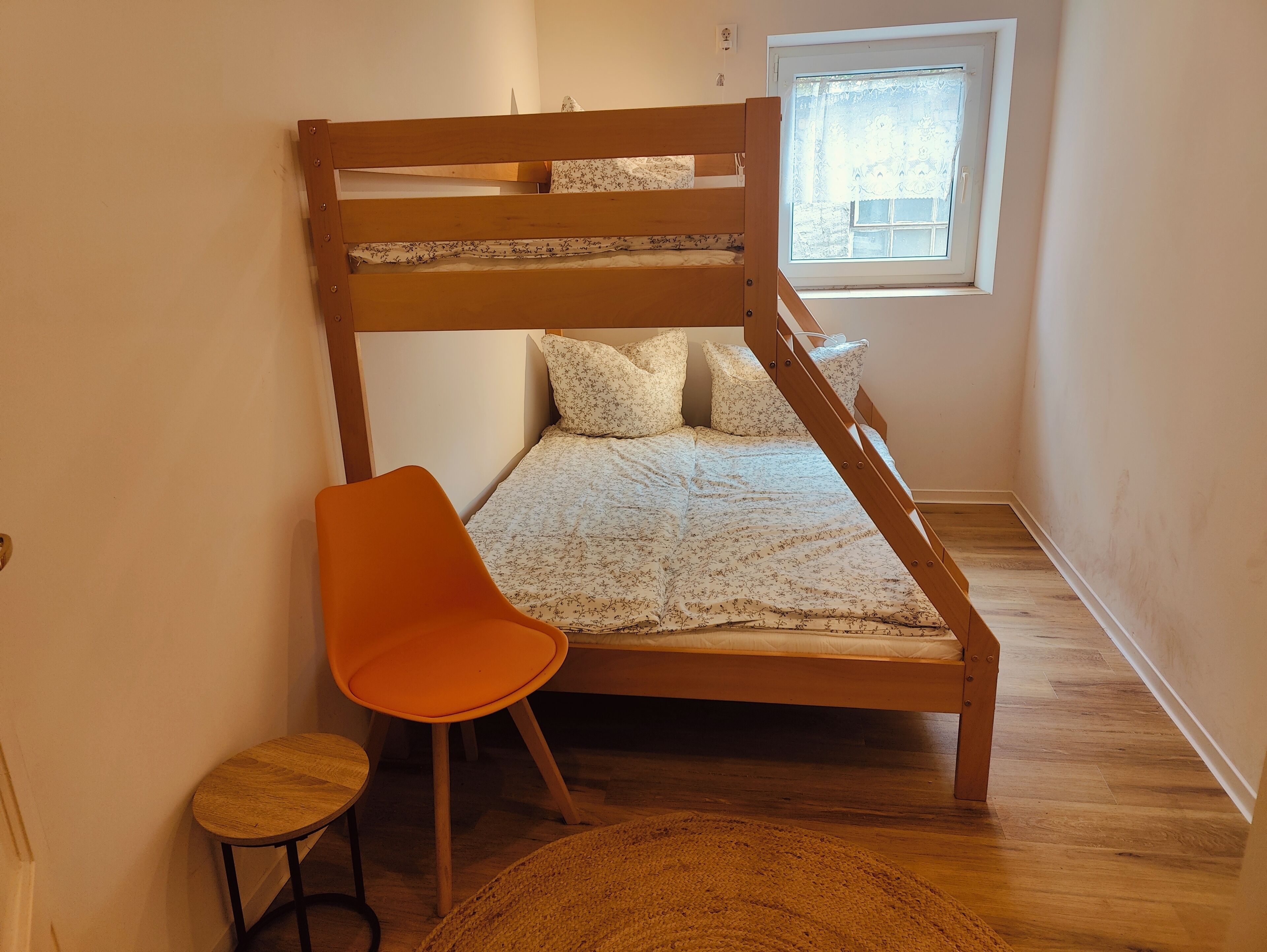 Una cama litera con estructura de madera, una pequeña silla naranja, un taburete de madera redondo y una ventana con vistas al exterior.
