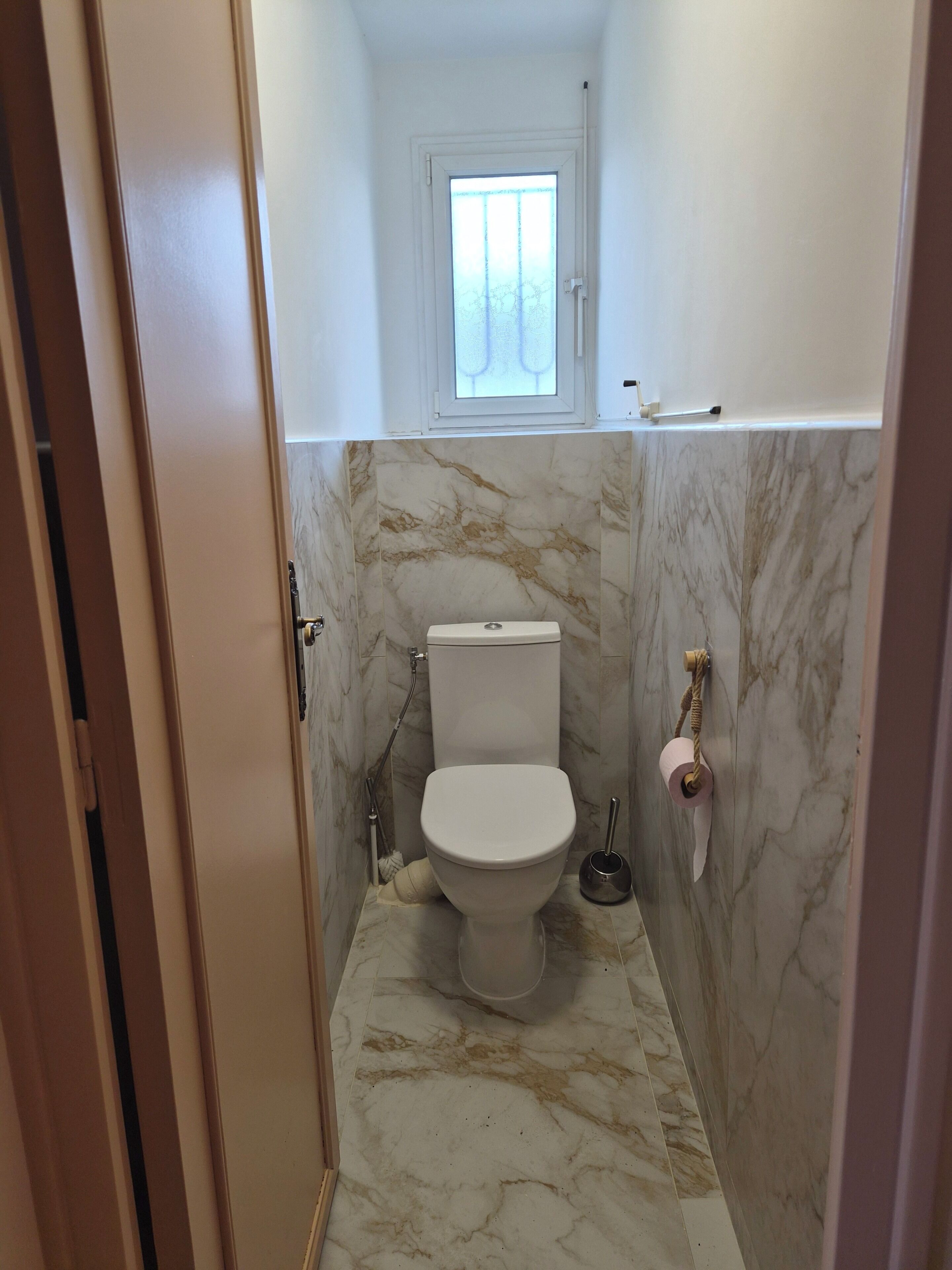 Baño compacto con inodoro, paredes de mármol, ventana y ducha.