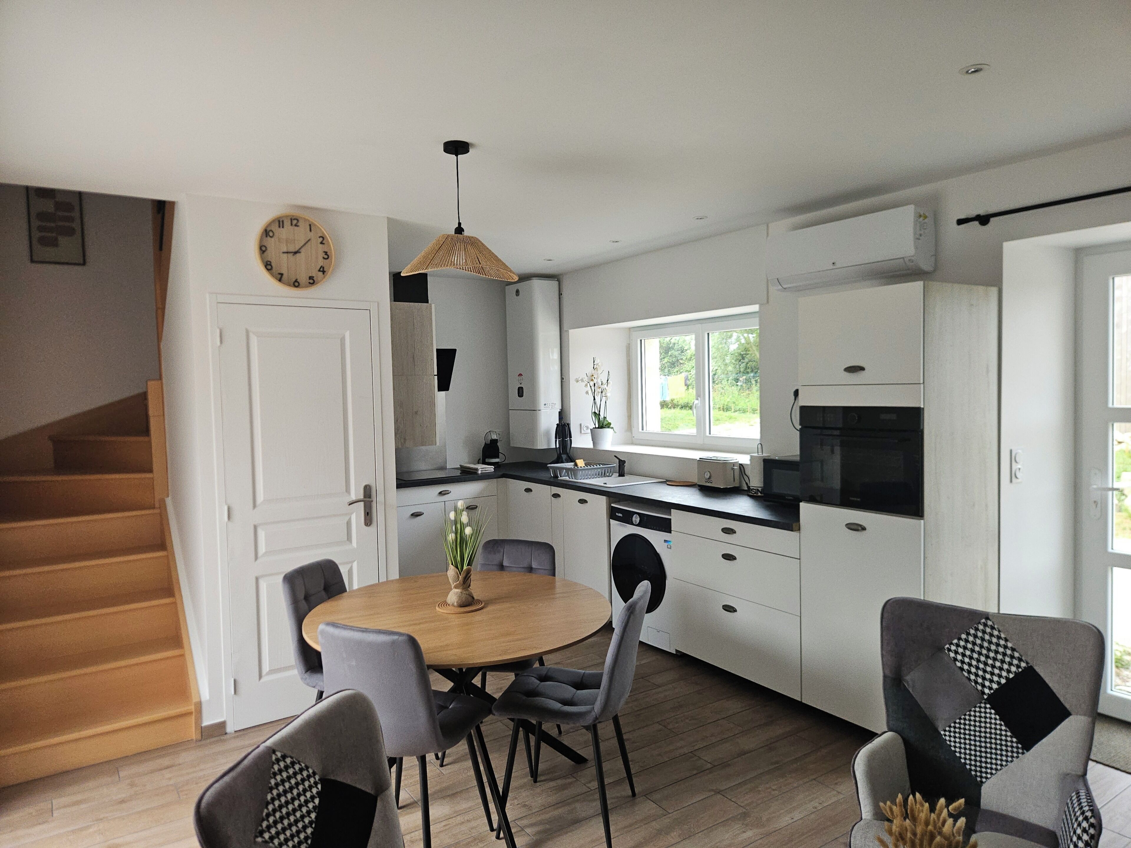 A modern kitchen with white cabinets, a dining table with chairs, a clock on the wall, and a staircase leading to another room.