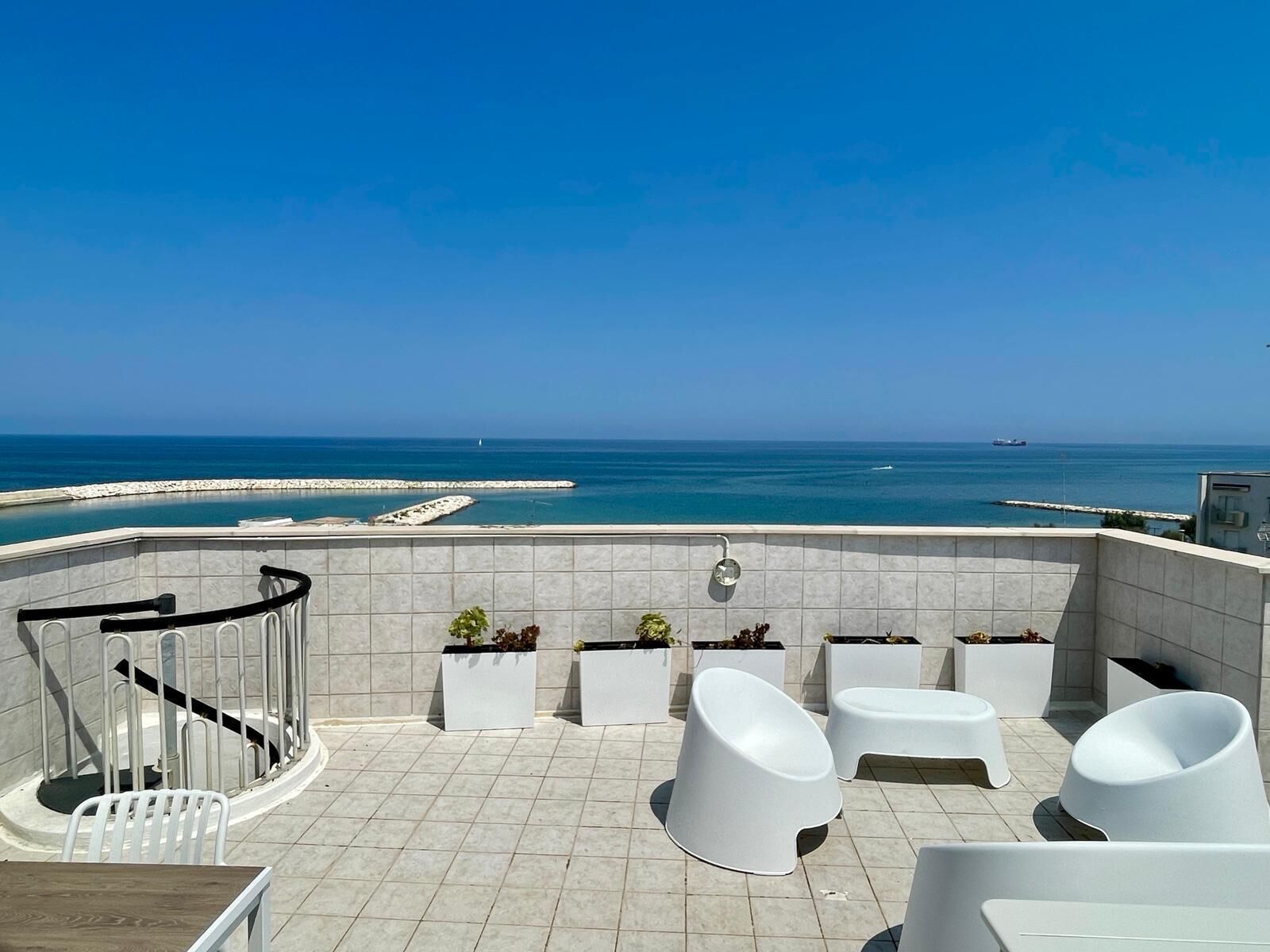A terrace with white chairs and planters, overlooking a coastal area with a clear sky.