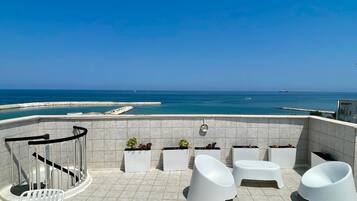 Una terraza con sillas blancas y macetas, con vistas a la costa y un cielo despejado.