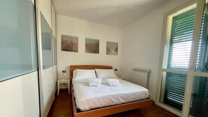 Un dormitorio con una cama de madera, ropa de cama blanca, una mesita de noche, un radiador y una ventana con persianas.