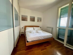 Un dormitorio con una cama de madera, ropa de cama blanca, una mesita de noche, un radiador y una ventana con persianas.